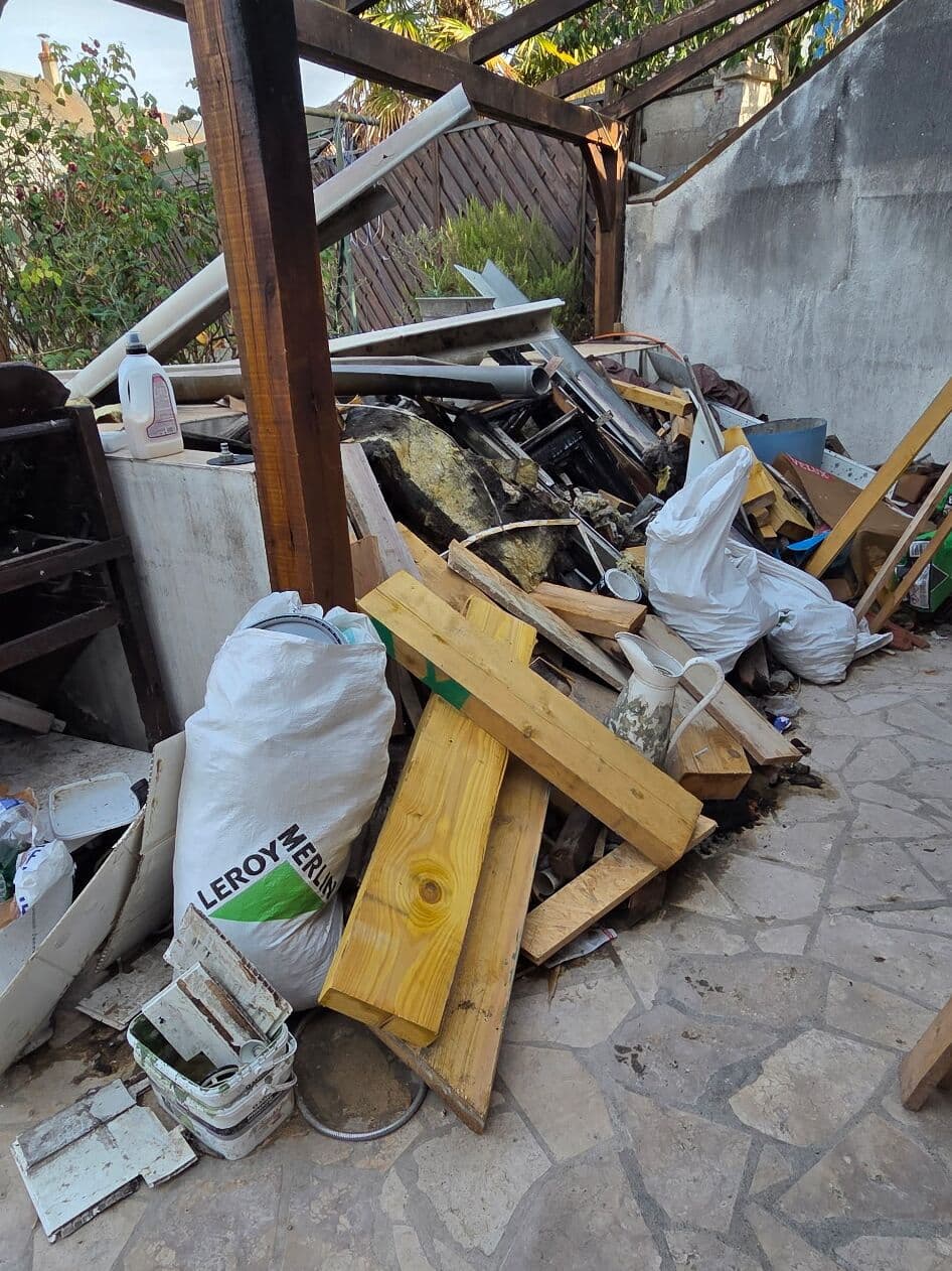 Planches, sacs de gravats, tuyaux et objets à jeter entassés sous une pergola, sur une terrasse dallée