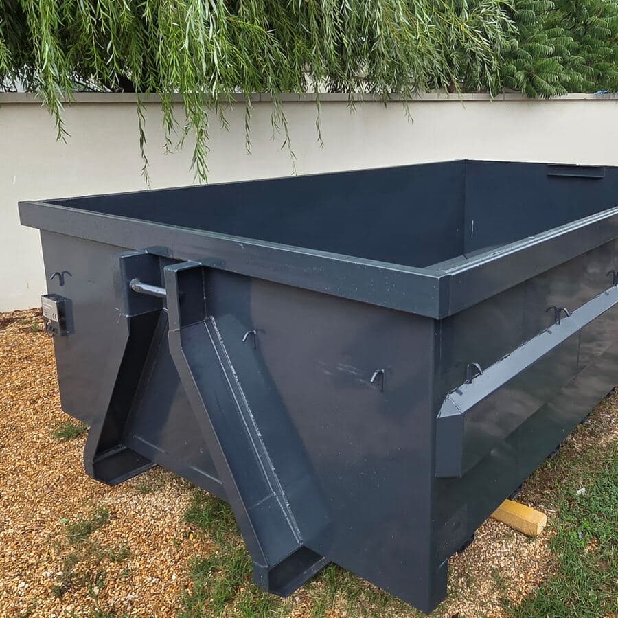 Benne métallique gris anthracite posée sur du gravier, au pied d’un mur, sous les branches d’un saule